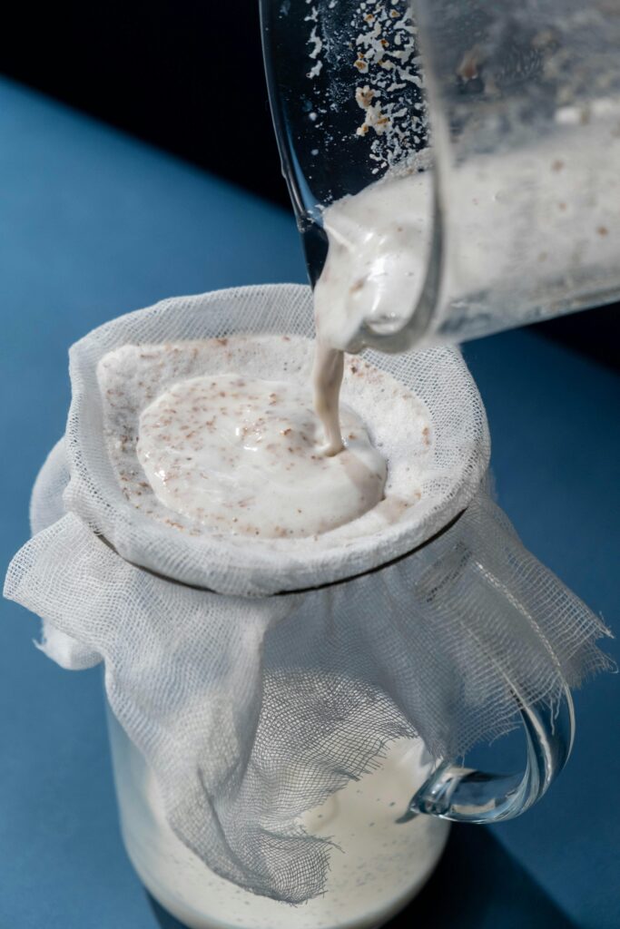 Straining homemade oat milk through cheesecloth into a glass for a smooth and creamy texture. DIY oat milk preparation process.